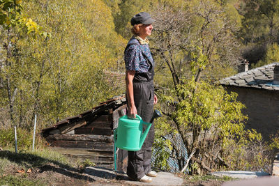 Side view of woman holding watering can while standing against trees