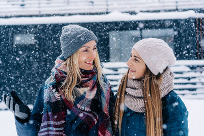 Portrait of two women in hats and jackets standing under the snow and looking at each other smiling.