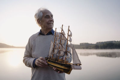 Smiling senior man holding toy boat while standing against lake