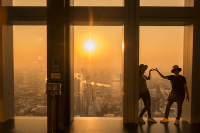People standing by window in city during sunset