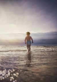 Rear view of child standing at beach against sky during sunset