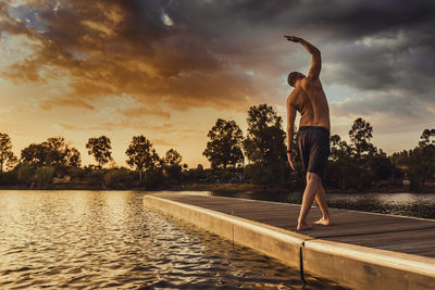 Man exercising by lake against sky during sunset