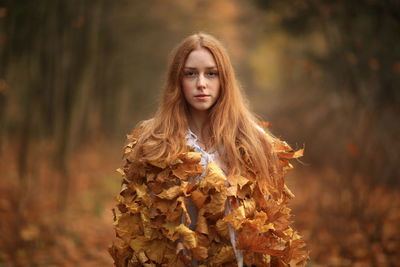 Portrait of young woman covered with leaves standing at forest during autumn