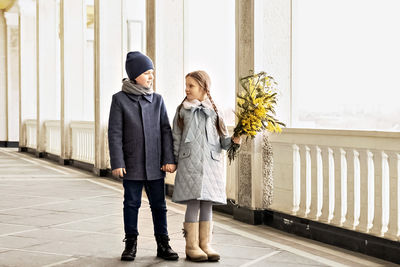 A couple of a boy and a girl of schoolchildren in spring coats with a bouquet of mimosa in  hands 