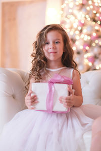 A beatiful girl with curly hair in white dress is holding of christmas box gift and sitting near 