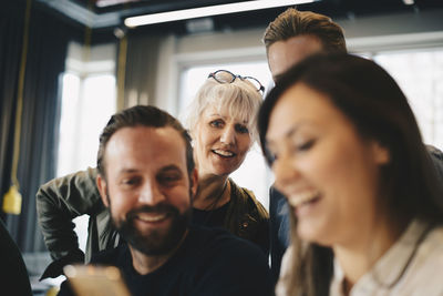 Happy businesswoman looking at colleagues using smart phone in office