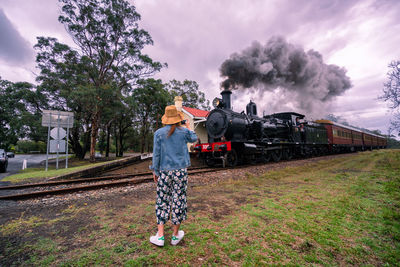 Rear view of woman standing by steam train on railroad track against sky