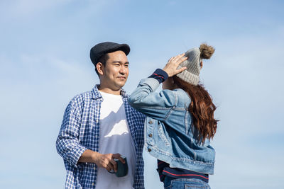 Man adjusting knit hat of woman while standing against sky