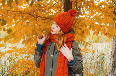 Young woman stands under tree with yellow leaves. dressed in trendy orange knitted hat and scarf. 