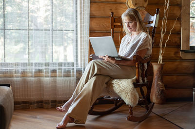  blonde woman in beige home clothes sits in a rocking chair with a laptop in her hands 