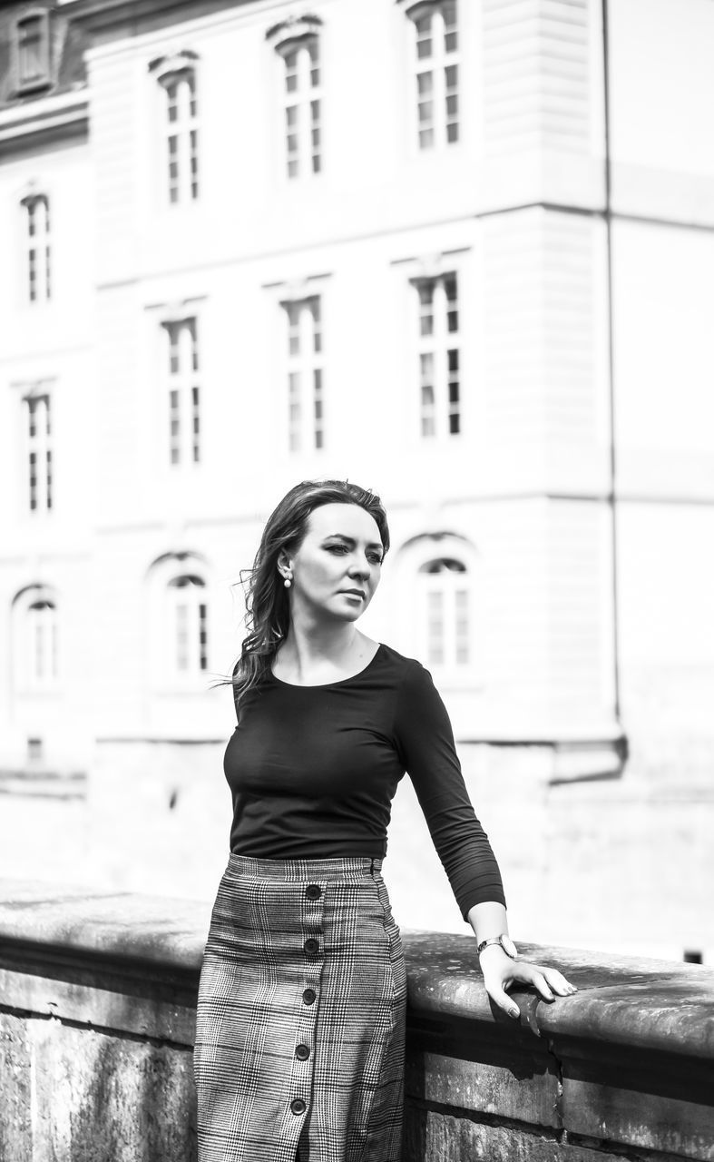 FULL LENGTH OF YOUNG WOMAN STANDING AGAINST BUILDING