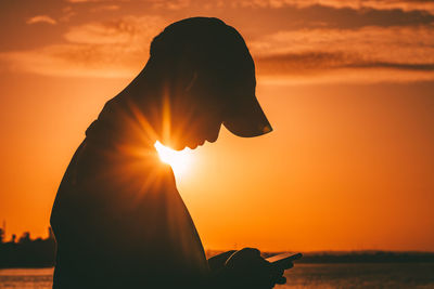 Silhouette of a young man standing at ocean using mobile phone against sunset