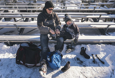 High angle view of father tying son's shoelace while sitting on bench during winter