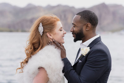 Side view of happy newlywed couple standing face to face against lake