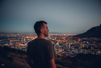 Rear view of man by illuminated cityscape against clear sky during dusk