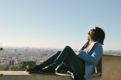 Woman sitting on retaining wall against clear sky