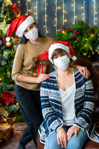 Adult mother and daughter near the christmas tree wearing medical masks. in caps of santa claus