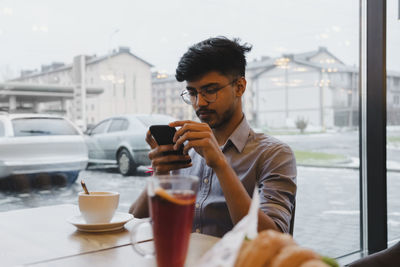 Young man using smart phone sitting by window in cafe