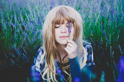 Beautiful young woman holding lavender flowers over eye while sitting in lavender field