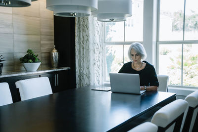 Businesswoman working on laptop computer while sitting against window in office