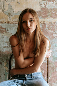 Portrait of shirtless young woman sitting on chair against wall