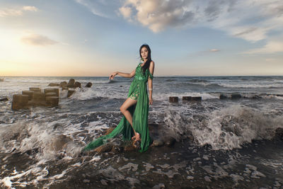 Portrait of young woman standing on beach against sky during sunset