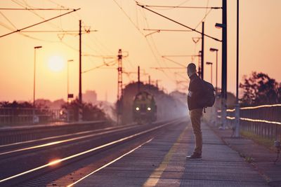 Side view of man standing at railroad station against sky during sunset