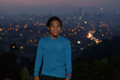 Portrait of smiling man standing against illuminated city at dusk