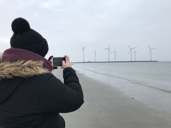 Rear view of man photographing windmills on mobile phone at beach