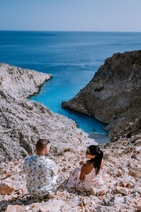 Rear view of couple sitting on rock formation by sea against sky