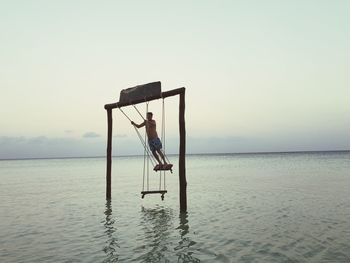 Shirtless man on swing over sea against clear sky