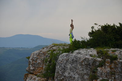 Wife jumping on rock by mountain against sky