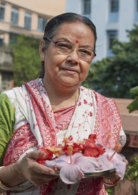 An aged bengali woman offering prayer to sun god, holding bouquet of hibiscus flowers in tray.