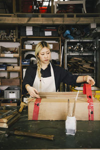 Young female entrepreneur packing cardboard box at workbench in workshop