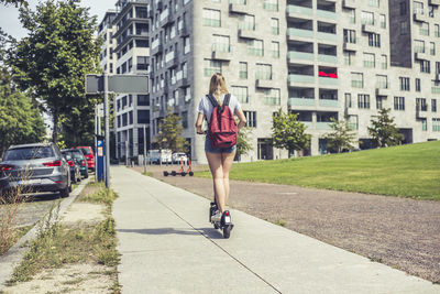 Back view of young woman with backpack riding e-scooter on pavement, berlin, germany