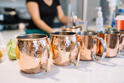 Close-up of cups arranged on table with woman in background at kitchen