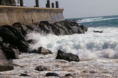 Waves splashing on rocks at shore against sky