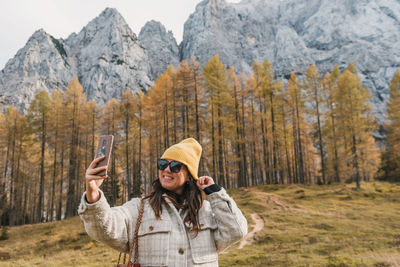 Portrait of happy young woman taking a selfie in front of golden larch trees under mountains