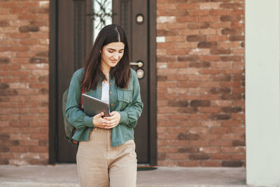 Young woman using phone while standing against brick wall