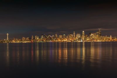 Illuminated buildings by sea against sky at night