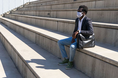 African american male entrepreneur wearing medical mask relaxing on stone stairs in city during coronavirus and looking away