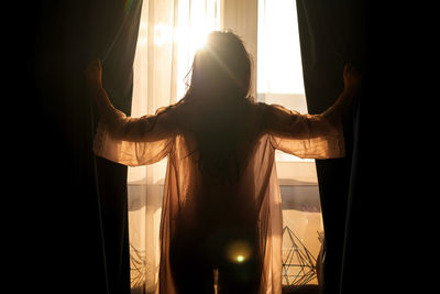 Silhouette of young woman standing near window open curtain in the morning of new day.