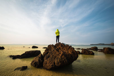 Rear view of young woman standing on rock in sea against sky