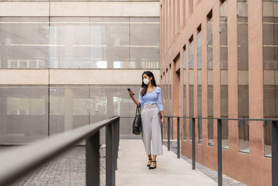 Businesswoman in protective face mask using smart phone while walking outside office building during coronavirus outbreak