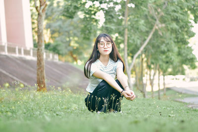 Portrait of young woman crouching on grass against tree at park