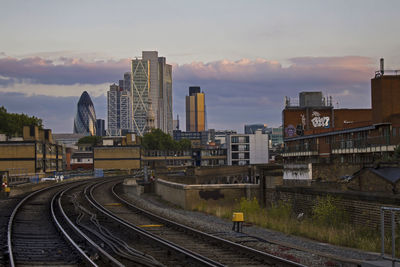 Railroad tracks by modern buildings against sky during sunset