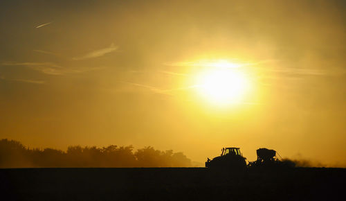 Silhouette of a tractor sowing seeds in a field in a cloud of dust against the background.