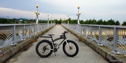 Bicycle parked by railing on footbridge against sky