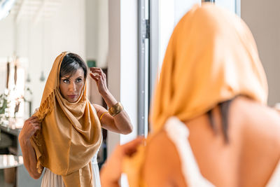 Attractive muslim female in traditional yellow headwear looking at reflection in mirror while standing in light spacious room at home