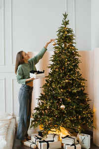 Young woman in home clothes decorates a christmas tree with toys in a cozy house